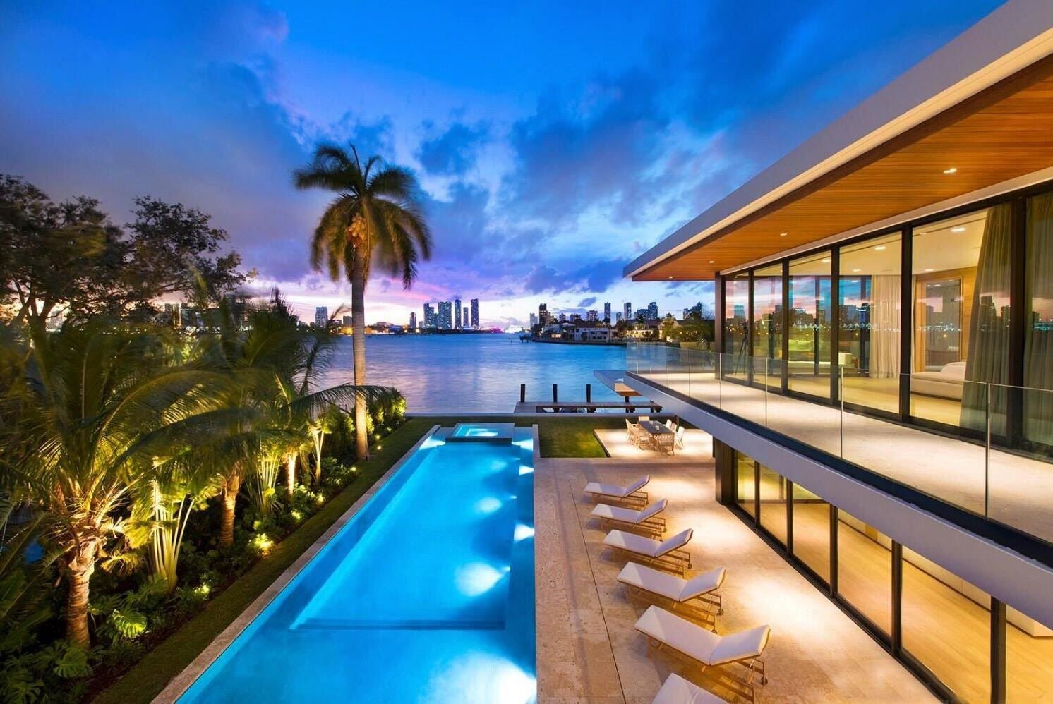 A stunning, modern waterfront mansion in Miami Beach at sunset, representing a billionaire's luxury purchase with yacht and palm trees