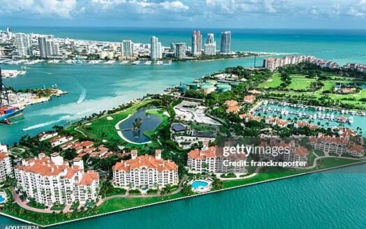 Aerial view of Fisher Island, Miami, Florida, featuring luxury condos, lush greenery, and marina against turquoise waters.