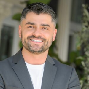 Smiling man in a gray blazer stands outdoors, with soft focus greenery in the background.